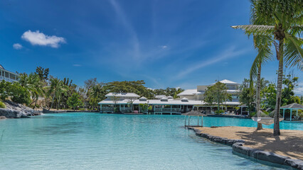 Recreation area by the swimming resort pool. The hammock is suspended from the trunks of palm trees on a small island. Canopy over aquamarine water.Cottages with terraces, deck chairs on the far shore