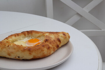 Fresh Adjarian style khachapuri with cheese and yellow yolk of an egg on round plate on white kitchen table near chair. Concept of lunch in cafe, national Georgian cuisine, eat. Horizontal food photo
