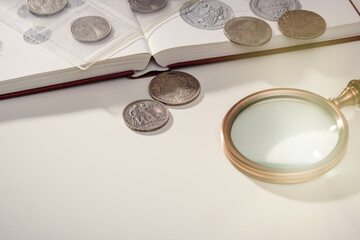 Numismatics. Ancient collectible coins and magnifying glass on a white table. Light background.