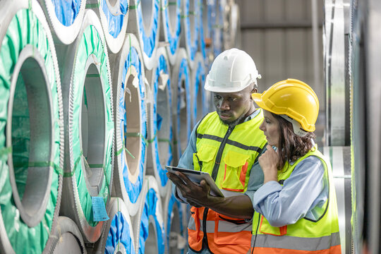 Two mechanical engineers in safety uniform from different nationalities working together checking steel stock in a factory. Culture diversity in a metal factory or warehouse. 
