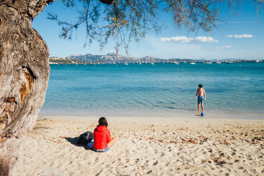 Famille &agrave; la plage. Jeune homme jouant au ballon sur une plage de sable fin. Femme assise regardant un enfant jouant au foot devant la mer m&eacute;diterran&eacute;e &agrave; Majorque, Bal&eacute;ares