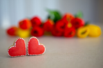 Heart-shaped cookies with tulip bouquet background. Two red heart-shaped cookies with white icing are displayed on a tabletop, with a blurred bouquet of red and yellow tulips behind them.