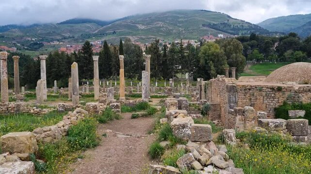 Panoramic view of Djemila village, surrounding hills, an ancient Roman bathhouse, and ruins with walls and columns, captured on a cloudy day