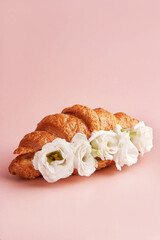 A minimalist composition featuring a croissant topped with white flowers against a soft pink background. A creative and unique take on a classic pastry
