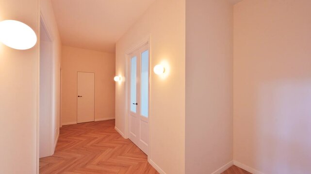 Minimalist hallway with warm beige walls, herringbone wooden flooring, white doors, and modern wall sconces casting soft glow.
