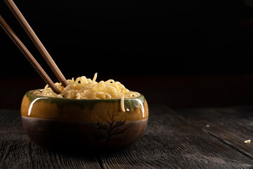 Steaming hot noodles being lifted from a  bowl by a pair of wooden chopsticks. The golden-yellow noodles cascade down, releasing wisps of steam that curl. Instant noodles on dark background. Food conc