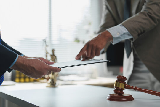 Two lawyers are discussing and reviewing a contract on a clipboard, with a gavel and scales of justice in the foreground, symbolizing legal proceedings and justice