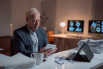 Focused senior businessman is working late in office using calculator, with computer screens showing charts and graphs in background, surrounded by paperwork and digital tablet
