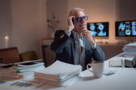 Senior businessman working late at night in his office, he is sitting at desk and talking on mobile phone while checking graphs on computer screens