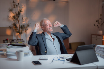 Senior manager raising arms and celebrating success while sitting at desk in office at night, working late with tablet, keyboard, paperwork and smartphone