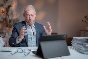 Senior businessman sitting at his desk in office at night having a video conference on a tablet looking surprised and gesturing with his hands