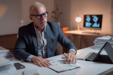 Senior business executive working late, reviewing financial data and analyzing charts and graphs at his desk in a dimly lit office, demonstrating dedication and hard work