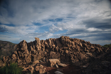 A rugged rock formation stands prominently amidst a breathtaking landscape, with clouds rolling in the sky above. The rocky surface reflects the natural beauty of the area.