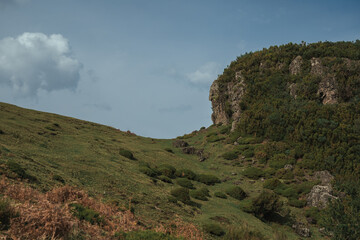 Lush green hills rise dramatically, bordered by rugged rock formations. The sky is partially cloudy, enhancing the natural beauty of this serene outdoors setting.