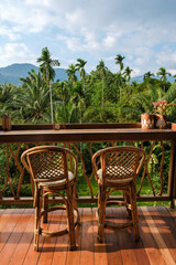 Obraz premium Two elegant bamboo chairs sit on a wooden deck, overlooking a vibrant tropical landscape filled with palm trees and mountains in the distance. This peaceful spot invites relaxation and tranquility.
