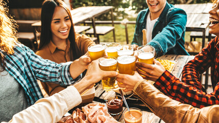 Group of multi ethnic friends having backyard dinner party together - Diverse young people sitting at bar table toasting beer glasses in brewery pub garden - Happy hour, lunch break and youth concept