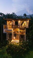 Nestled in lush greenery, this enchanting treehouse in Bali showcases warm, inviting lights as dusk falls. Guests enjoy serene moments on the deck, enveloped by natures beauty in the jungle rainforest