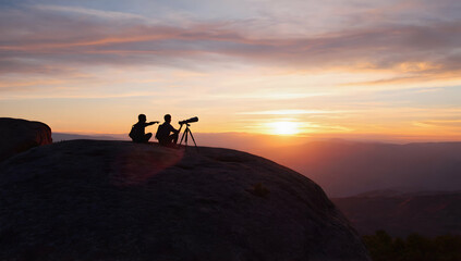Fototapeta premium Two people adjust a telescope atop a rocky outcrop at sunset