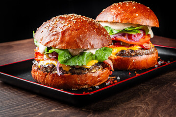 Two delicious gourmet burgers with sesame seed buns, stacked with fresh lettuce, tomatoes, red onions, cheddar cheese, and a creamy sauce, served on a black plate against a dark background.