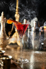 A martini is served in a martini glass with an orange slice on top. The glass is sitting on a table with a silver martini shaker and a silver cocktail glass. The scene is set in a bar or a restaurant