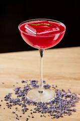 red cocktail in a coupe glass with a large ice cube stands on a table decorated with flowers