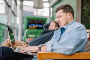 Obraz premium A focused man in a denim jacket working on a laptop in a lounge, with another man relaxing in the background. Modern indoor setting.
