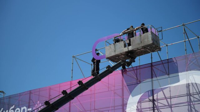  Specialists install street advertising in the cradles of an aerial platform or crane. Professional installers work Installation of a sign on the building. Bottom view