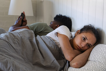 A sad woman lies in bed, looking away with a troubled expression, while her partner faces the other...
