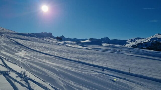 Picturesque shot from a ski lift overlooking the glistening white slopes and a race track with a few skiers. The sun shines over the snow-covered alpine mountains of Switzerland