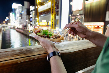 Fototapeta premium A tourist is taking a takoyaki ball with chopsticks, a popular street food in Osaka, Japan. It's a summer night, the lights of the billboards are reflecting on the canal water in the background