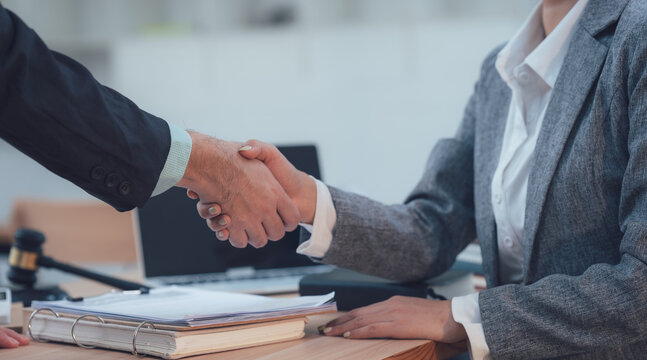 Sealing the Deal: Two business professionals in formal attire, engage in a firm handshake over a table laden with documents and legal paraphernalia.