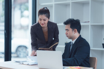 Business Collaboration: A focused business meeting scene. A confident female leader in a suit guides a male colleague while he works on a laptop in a modern office space.