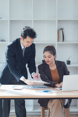 Focused Collaboration: Two business professionals, a man and a woman, intently review documents and data on a laptop, showcasing focused teamwork and dedication in a modern office setting. 