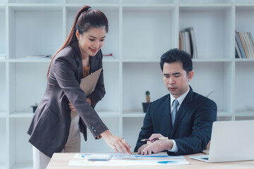 Business Collaboration: A focused business meeting unfolds as a female executive assists her male colleague, offering guidance and support. The scene is set in a modern, bright office space.