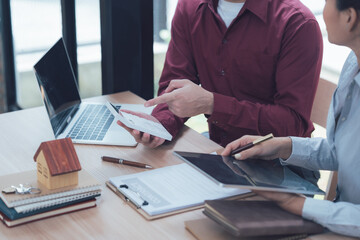 Real Estate Consultation: A real estate agent points to a tablet while discussing property details with a client, surrounded by paperwork, a laptop, and a miniature house model.