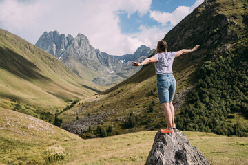 Naklejka premium A girl stands on a stone against the background of the Chaukhi mountain range near the village of Juta in Georgia. Mountains surround the girl on the stone. Mountain peaks of the North Caucasus