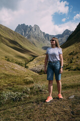 Naklejka premium A girl stands on a stone against the background of the Chaukhi mountain range near the village of Juta in Georgia. Mountains surround the girl on the stone. Mountain peaks of the North Caucasus