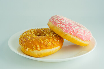 Image shows a simple arrangement of doughnuts on a white plate, featuring three distinct doughnuts one pink-glazed with possible nut toppings, another golden-brown plain or simply glazed, and a thir