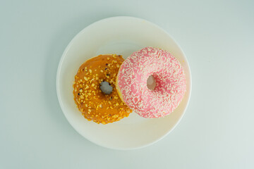 Two donuts on a white surface one yellow with scattered seeds or nuts, the other pink and sprinkled with white crunchy bits Ring-style, neutral background