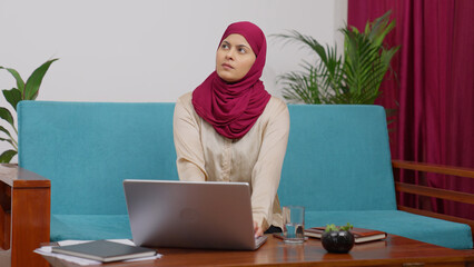 Muslim Woman in Hijab Scarf Sitting on Couch Working on Laptop and Thinking