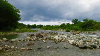 Bed of a shallow mountain river. View of a shallow river with rocky banks flowing through a valley among tropical trees on a cloudy day.
