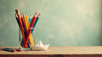 Obraz premium A Colorful Collection of Pencils in a Glass Holder with a Small Origami Boat on a Wooden Surface Against a Soft Teal Background