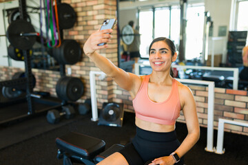 Fototapeta premium Fitness woman capturing post-workout selfie, smiling near gym equipment, radiating wellness and positive energy