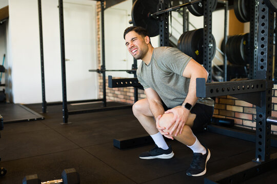 Muscular male grimacing from knee discomfort following strenuous gym training, revealing potential exercise-related physical strain