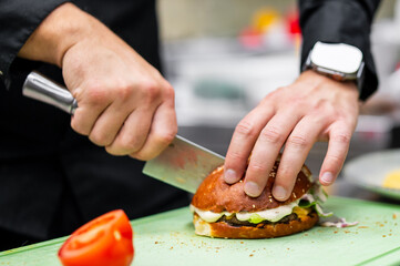 A chef's hands skillfully chop a freshly made sandwich on a green cutting board, with a sliced tomato nearby. The focus is on the preparation of a delicious meal.