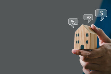 Hand holding miniature wooden house with financial symbols. Concepts of investment, real estate taxes, and property market insights.