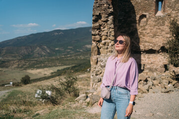 Fototapeta premium A tourist girl in a lilac blouse, blue jeans, and sunglasses against the background of ancient ruins. The place of power is Jvari in Georgia