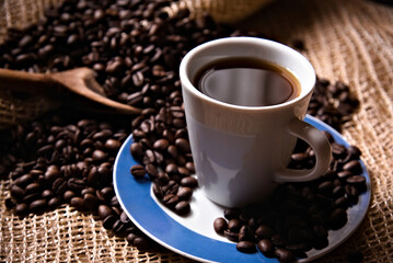 Freshly brewed coffee inside a coffee cup and roasted coffee beans on a rustic surface with a wooden spoon. Side view