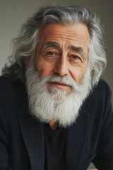 Portrait of a senior man with long gray hair and beard, wearing a dark jacket against a neutral gray background. The image is softly lit, focusing on