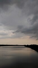 A serene dam with calm waters reflecting the cloudy sky, bordered by lush greenery and distant power poles, under an overcast atmosphere.